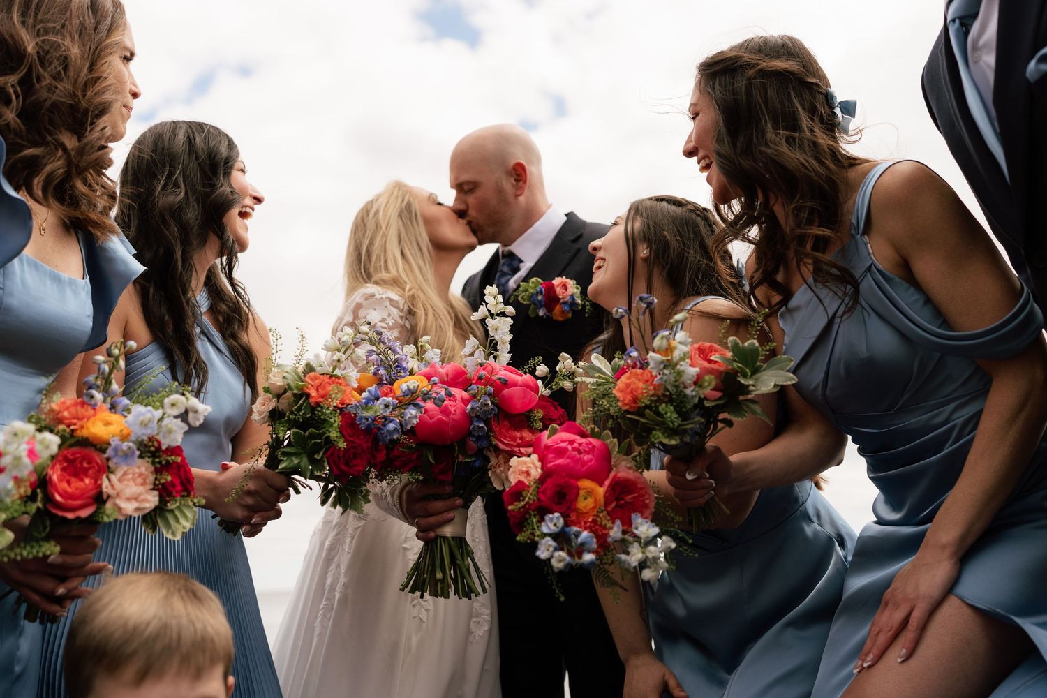 Bride and groom share a kiss surrounded by bridesmaids in blue dresses holding colorful bouquets at an outdoor wedding.
