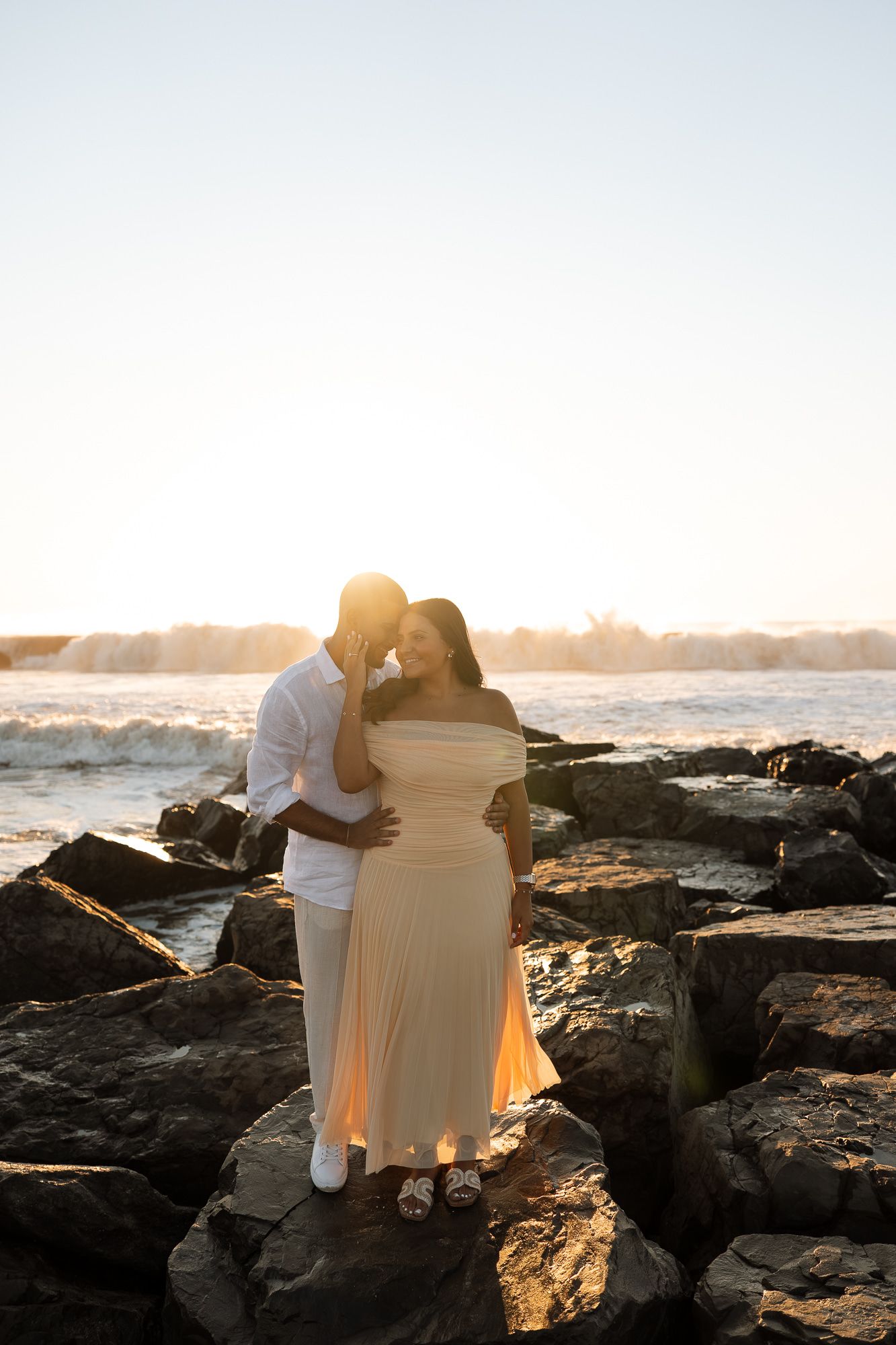 Couple embraces on rocky beach at sunset with golden light illuminating the coastline and ocean waves.