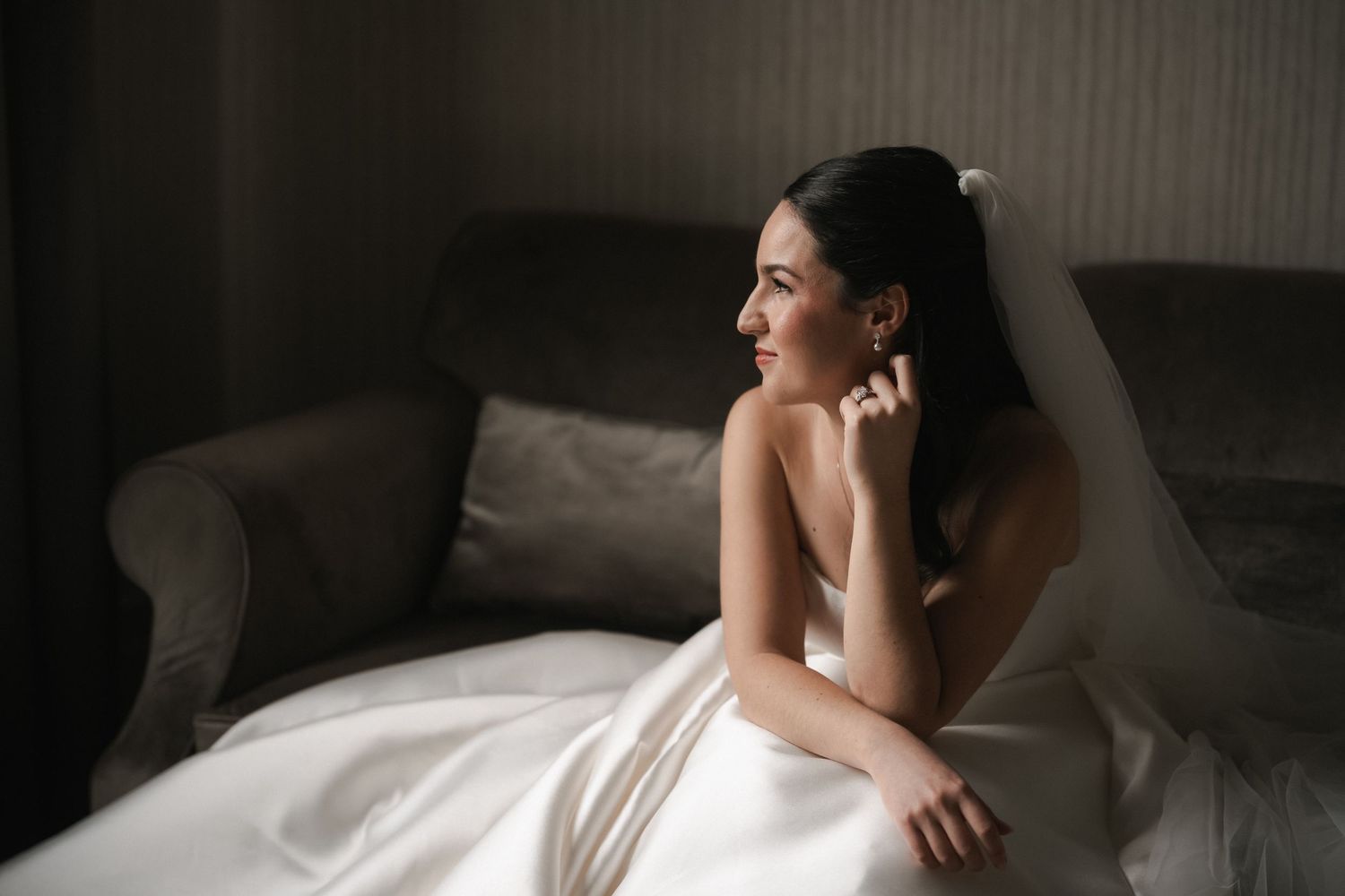 Elegant bride in white gown and veil sits on sofa, gazing thoughtfully toward window in soft natural light.
