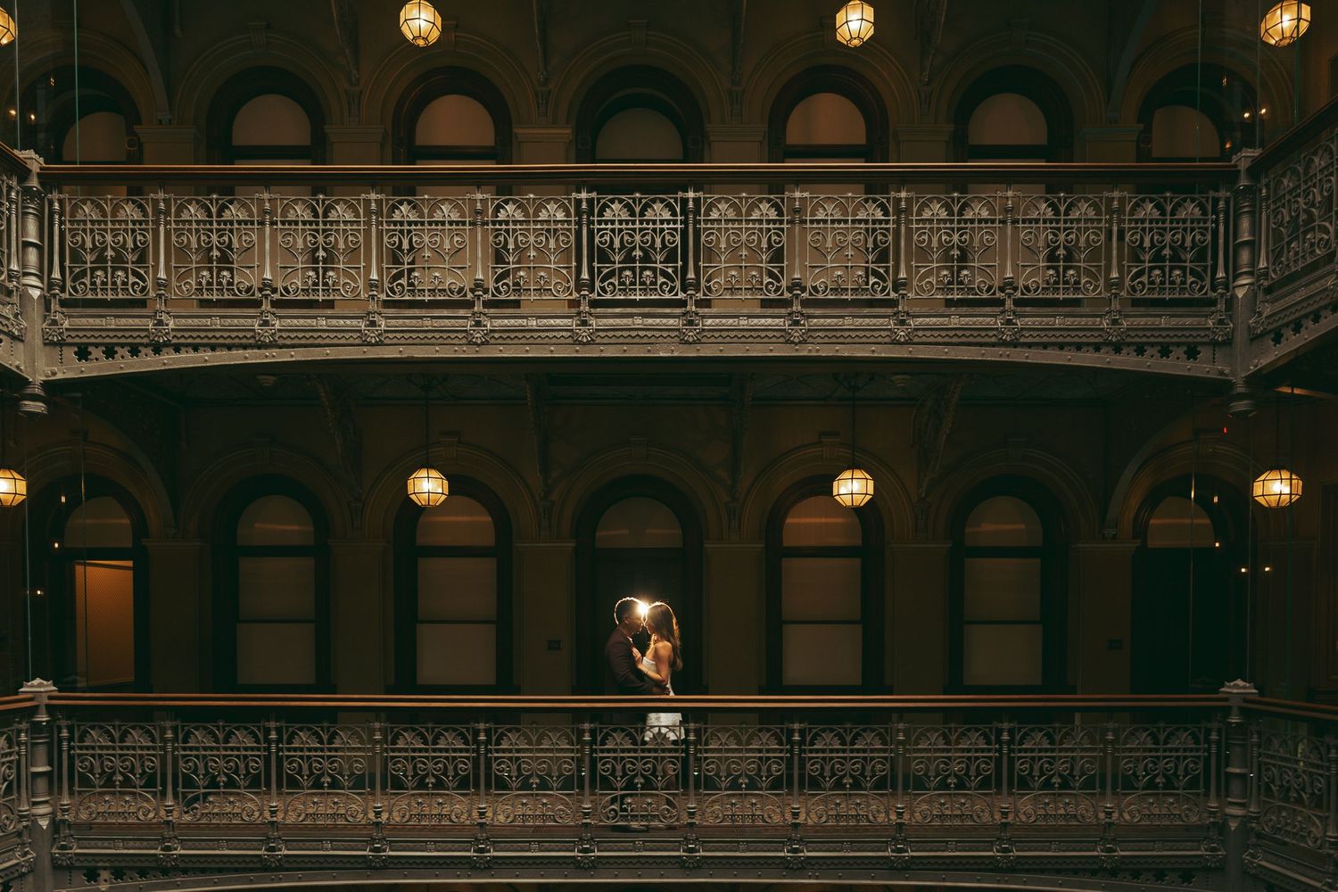 Couple embracing on ornate balcony of historic building with decorative iron railings and warm pendant lights.