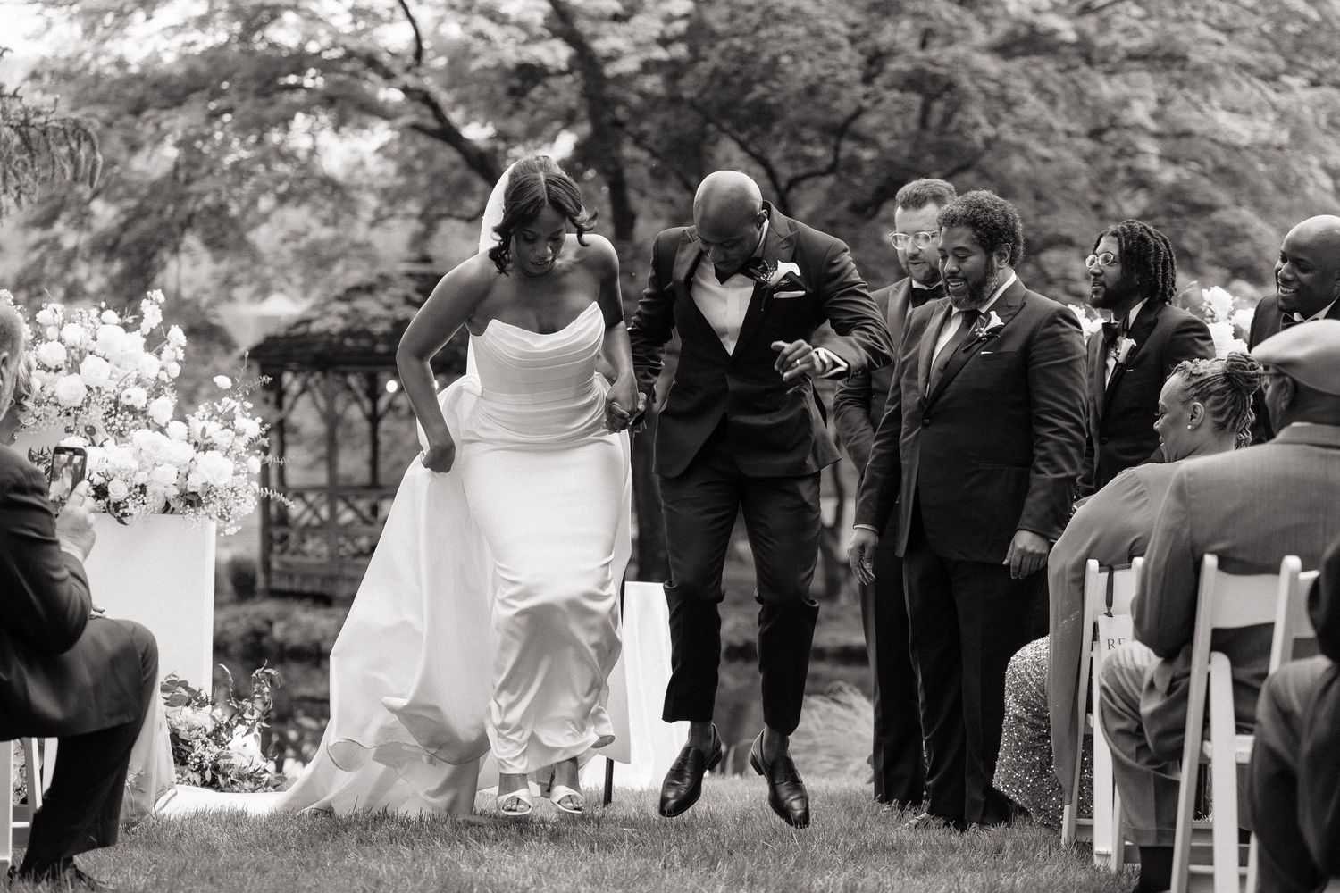 Joyful newlyweds jump together at outdoor wedding ceremony, groom leaping with excitement as bride holds dress, guests watching.
