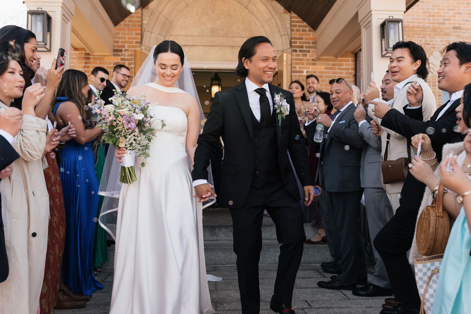 Bride and groom walk hand-in-hand past cheering guests outside a brick church after their wedding ceremony.