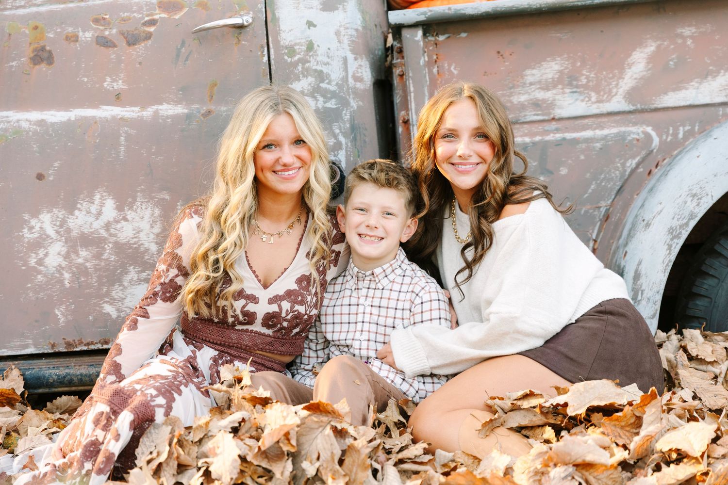 Three smiling children sit together in autumn leaves against a vintage rustic truck in a fall family portrait.