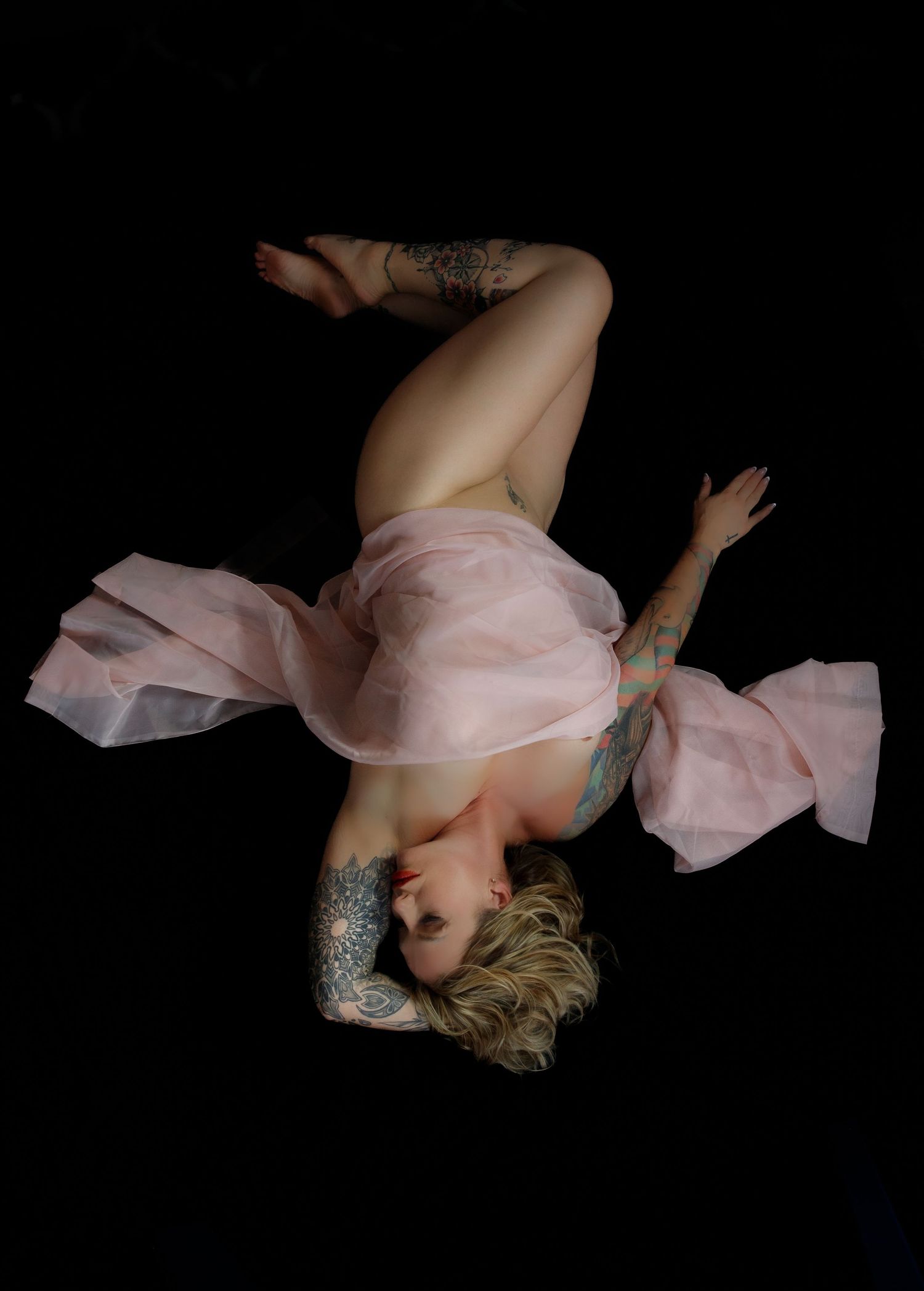 Figure in a soft pink flowing dress lies gracefully against dark background with visible tattoos and short hair.