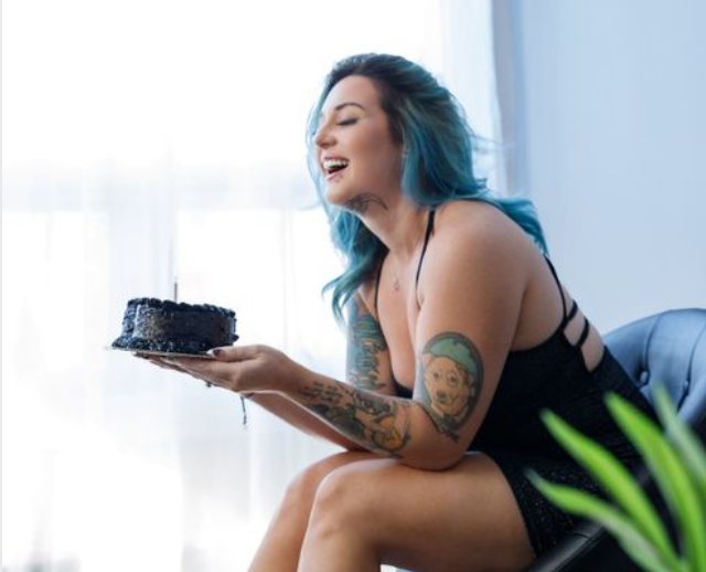 Woman with blue hair and tattoos laughing while holding a chocolate birthday cake indoors.