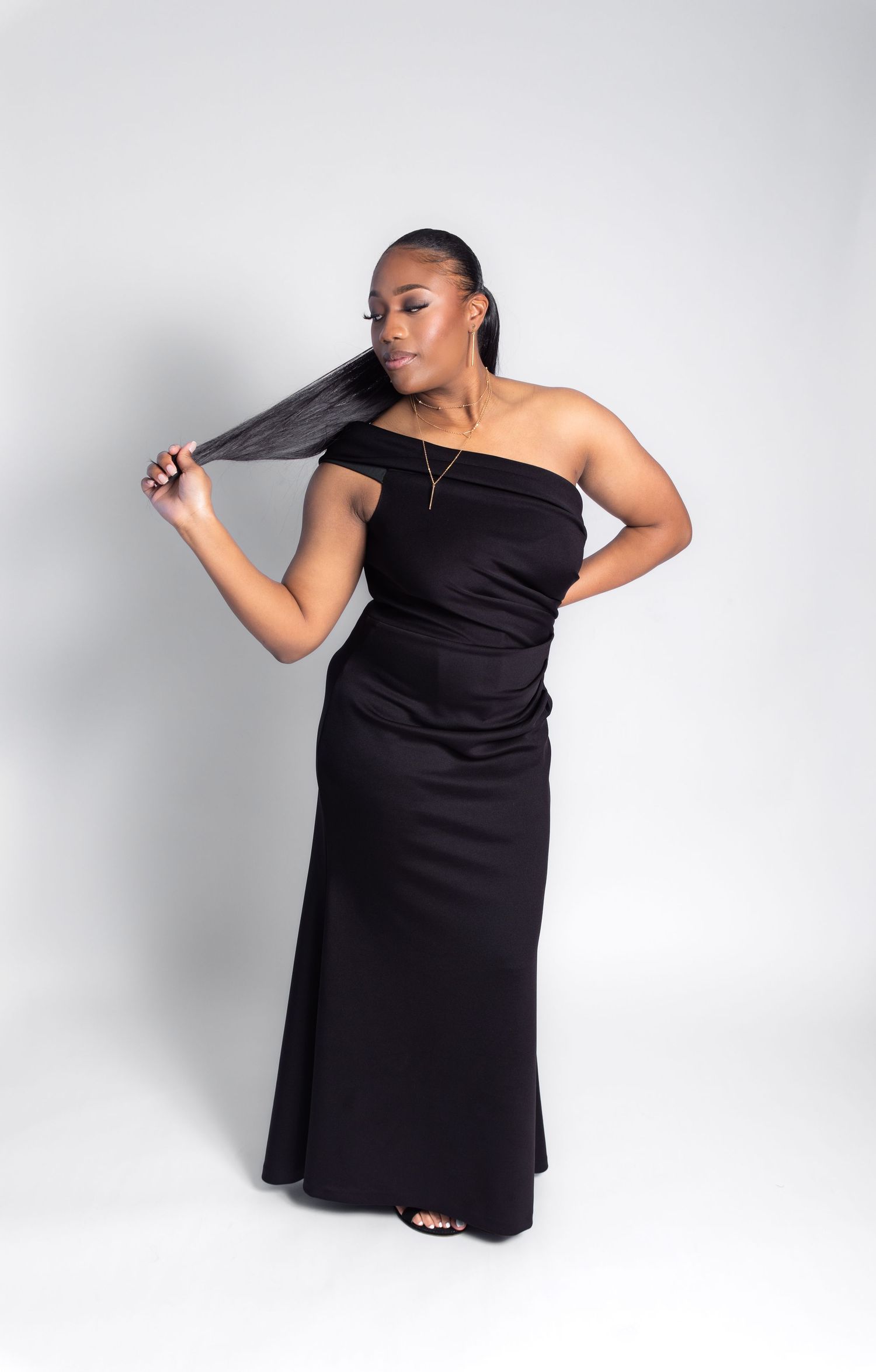 Woman in black one-shoulder maxi dress holding long hair extensions, posing against white background.