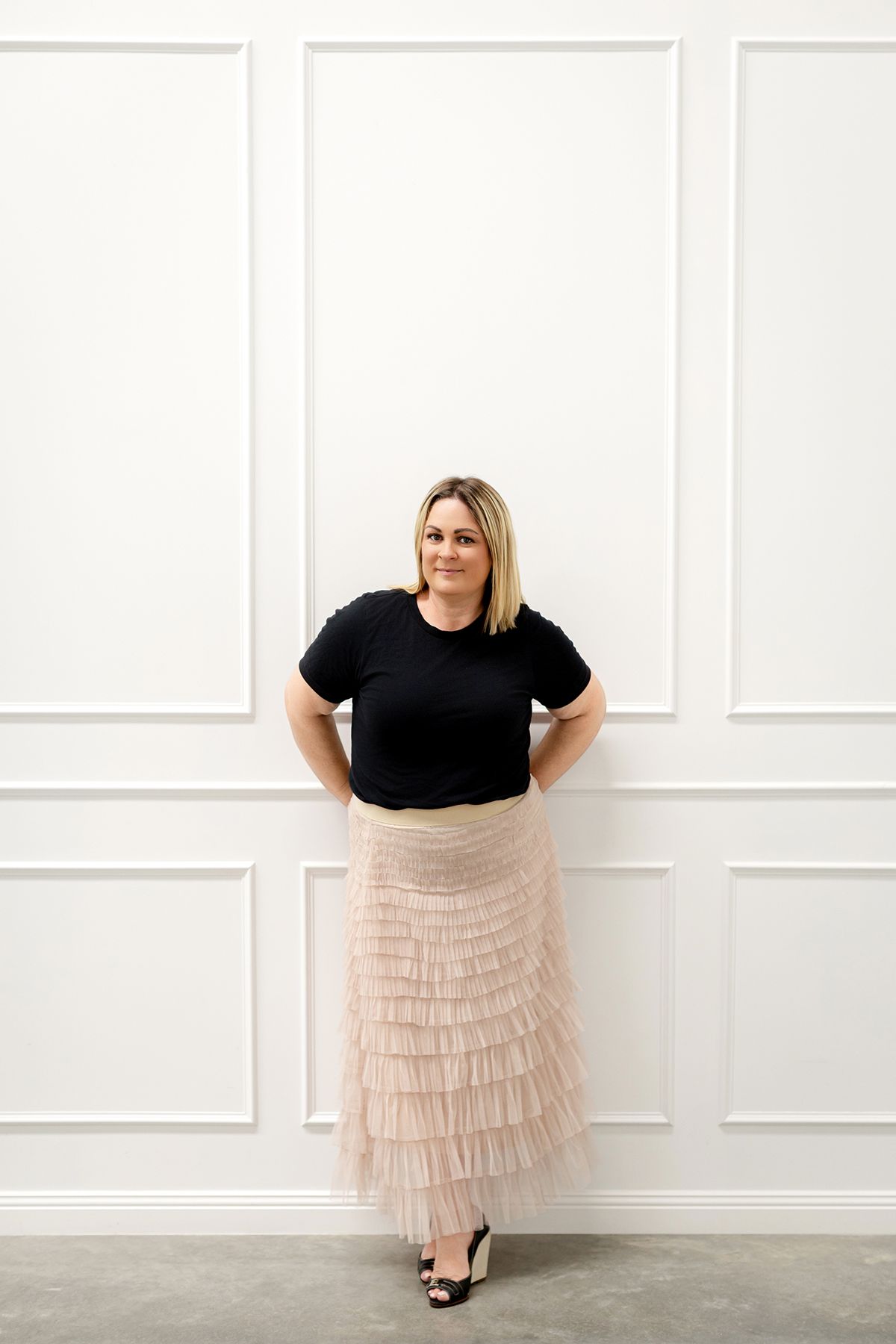 A person in a black top and tiered blush tulle skirt poses against a white paneled wall in four different stances.