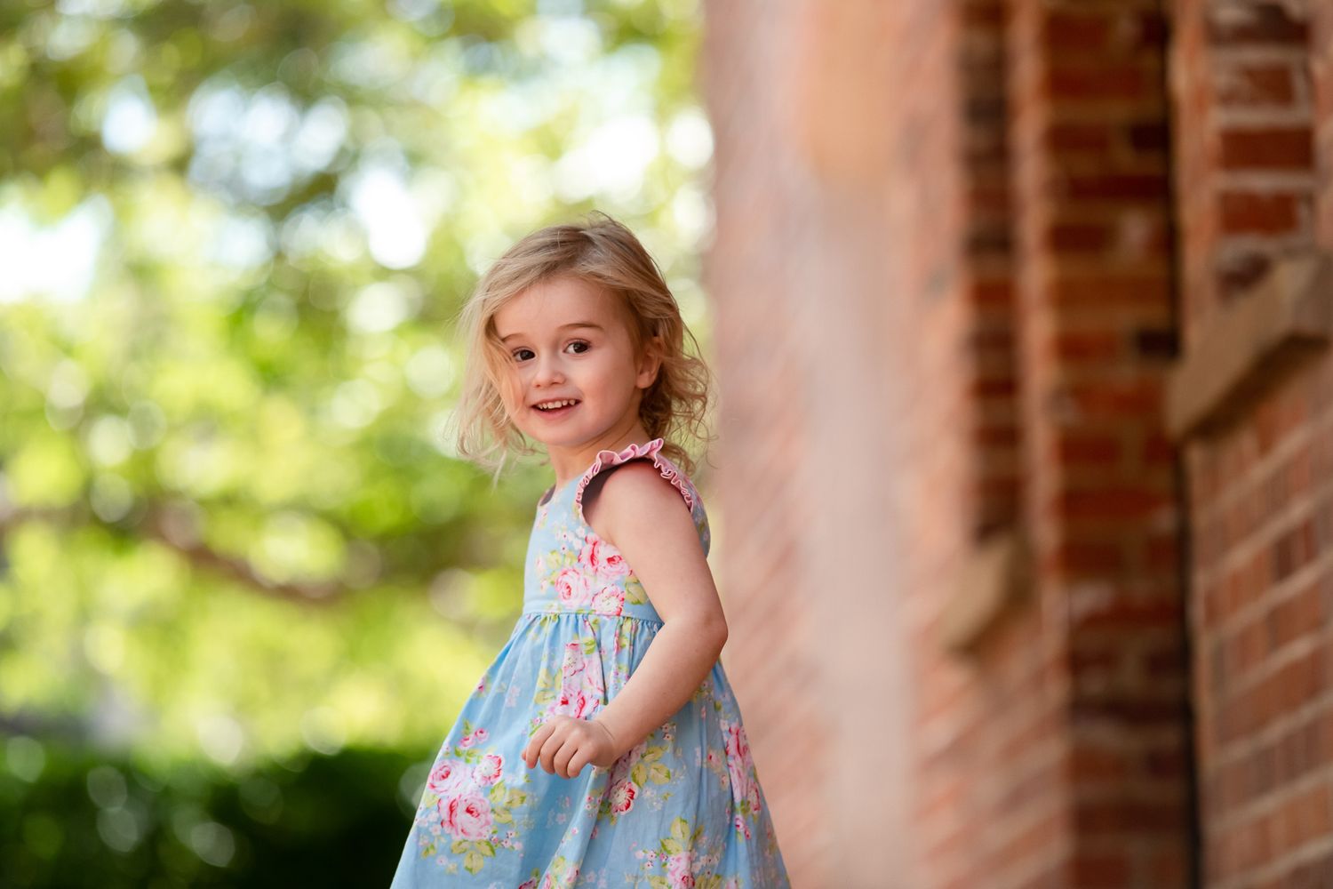 Young blonde girl in floral blue dress smiling beside brick wall with green trees in background.