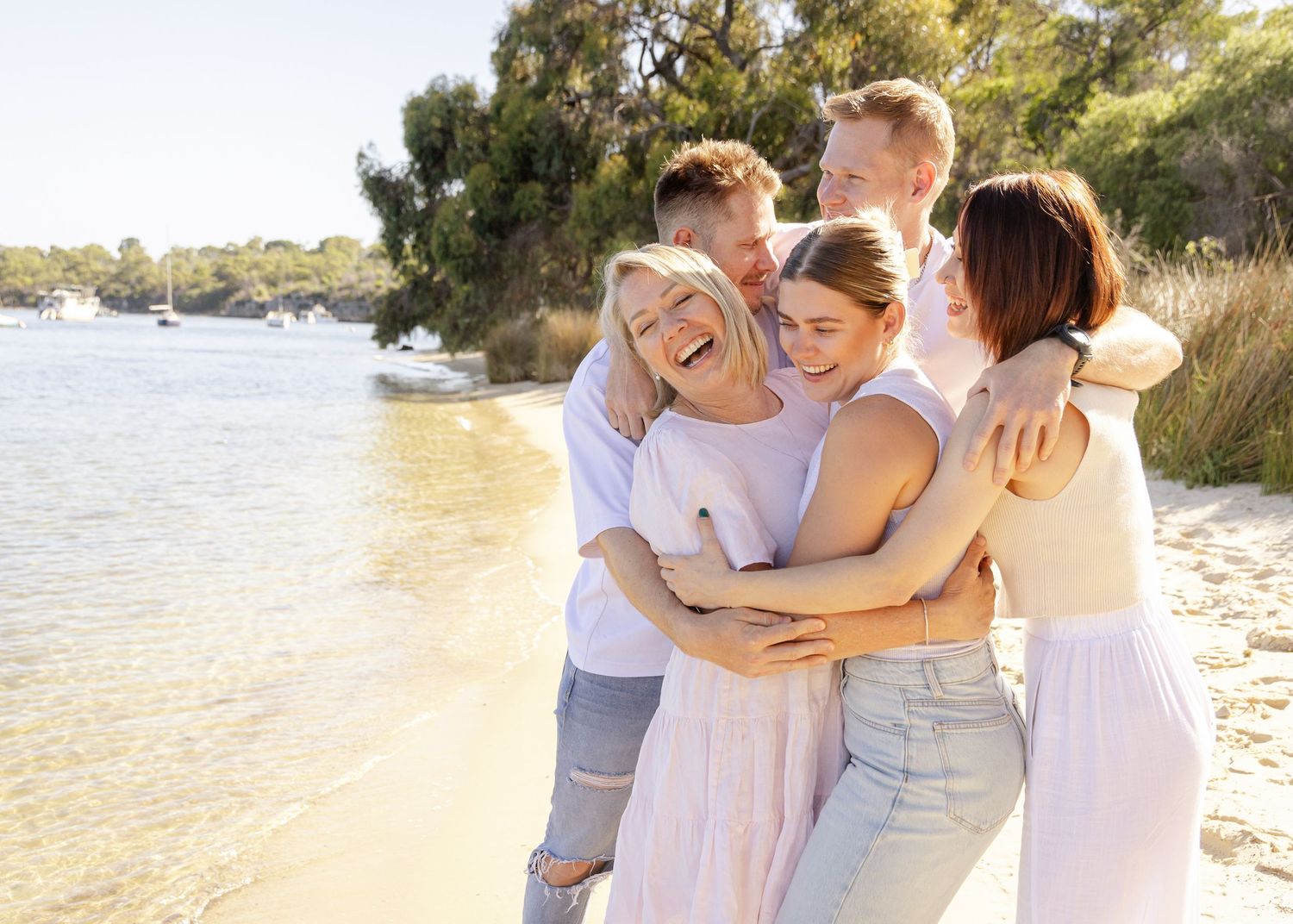 A group of people in white and neutral colored clothing embrace while walking along a sandy beach at sunset.