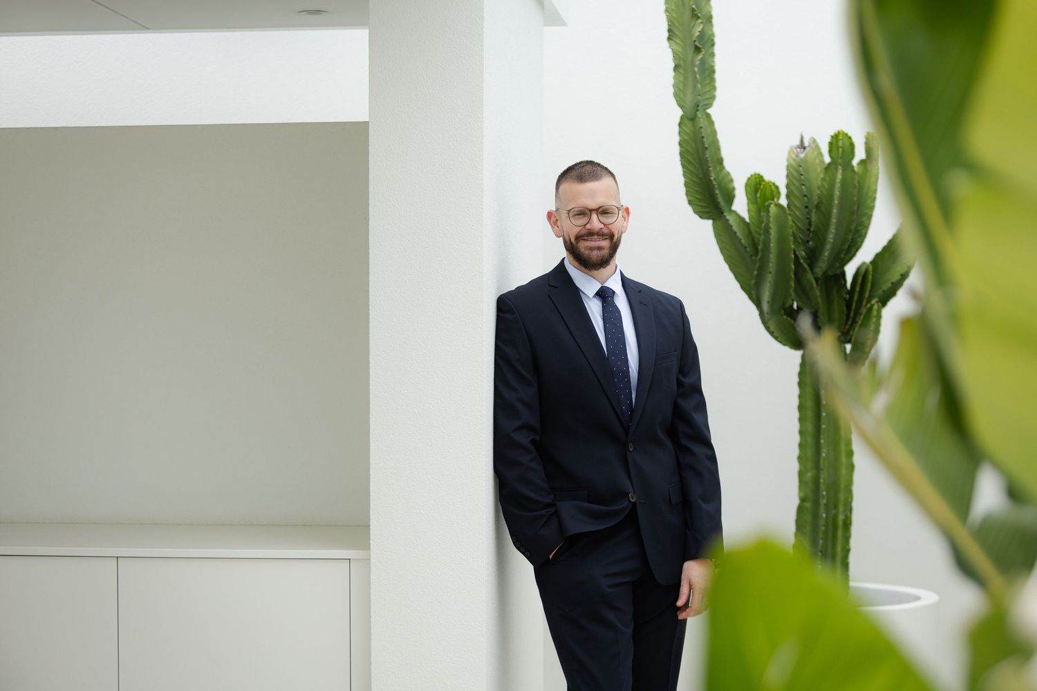 Professional wearing a dark suit poses against a white wall with green plants in soft focus at the edges.