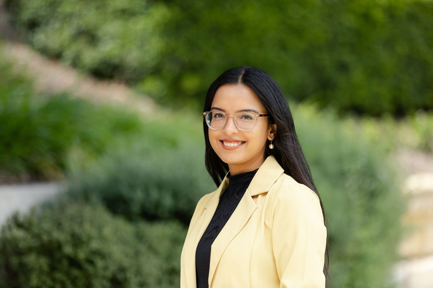 Professional portrait of someone in a yellow blazer and black top smiling outdoors with greenery in background.