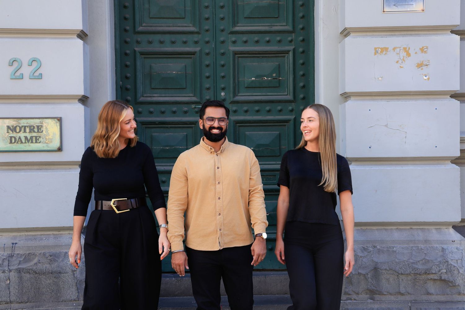 Three people smiling in front of a green door at Notre Dame building number 22.