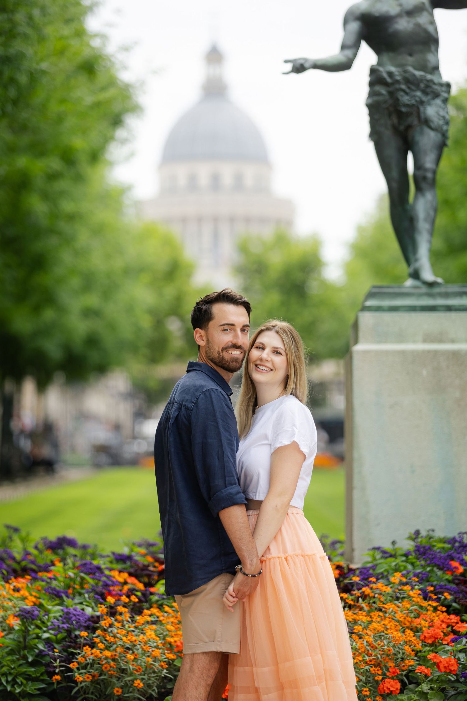 Couple embracing in a Parisian garden with colorful flowers, a bronze statue, and the Pantheon dome in the background.