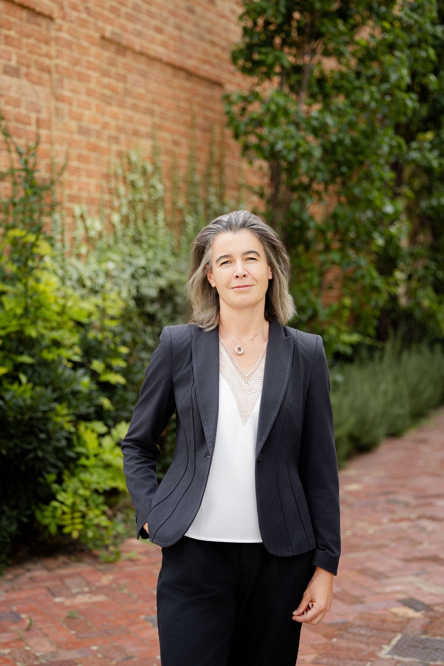 Professional woman in black suit with gray hair poses outdoors near brick wall and lush greenery.