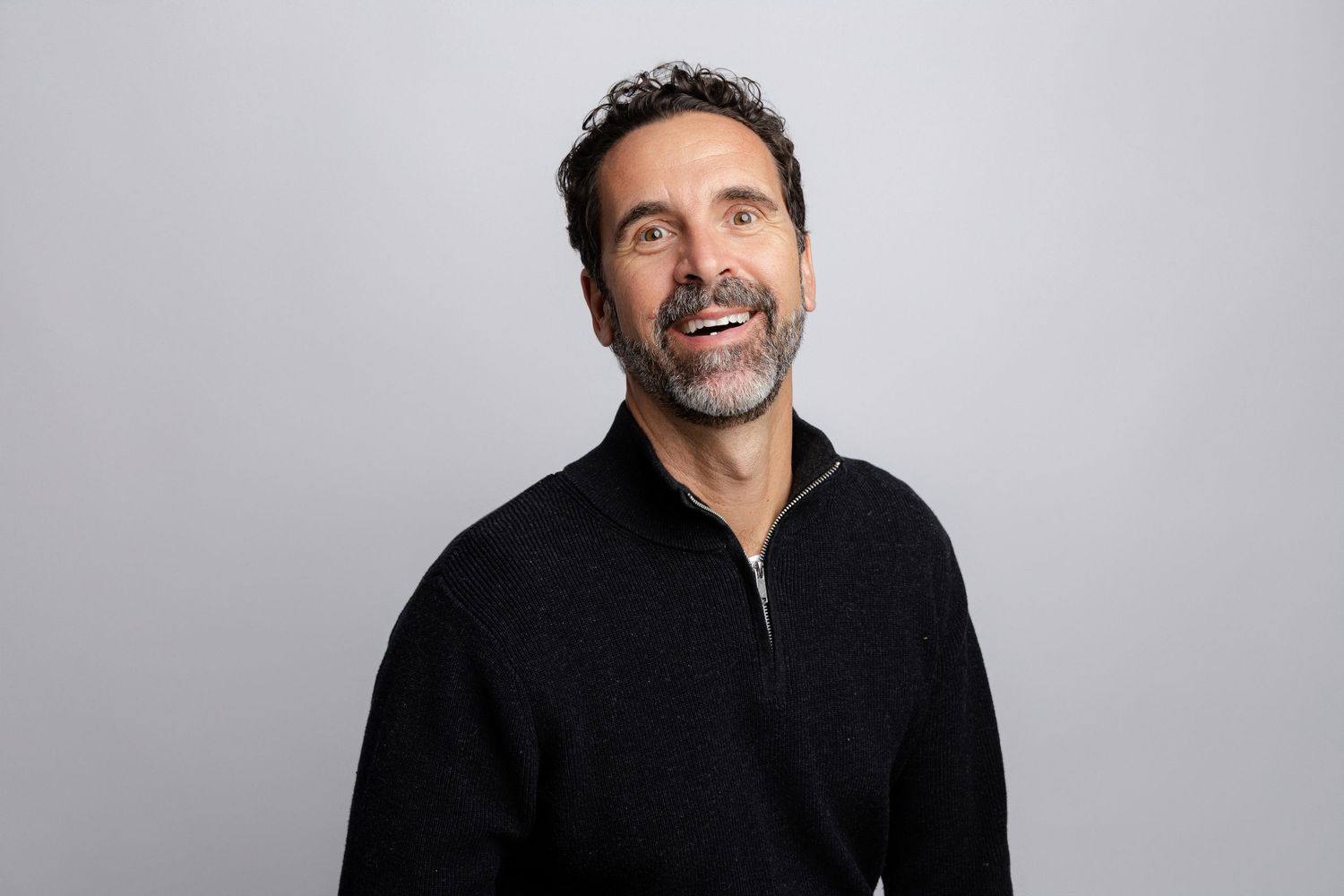 A person wearing a black zip-up sweater smiles warmly against a light gray background in a professional headshot.