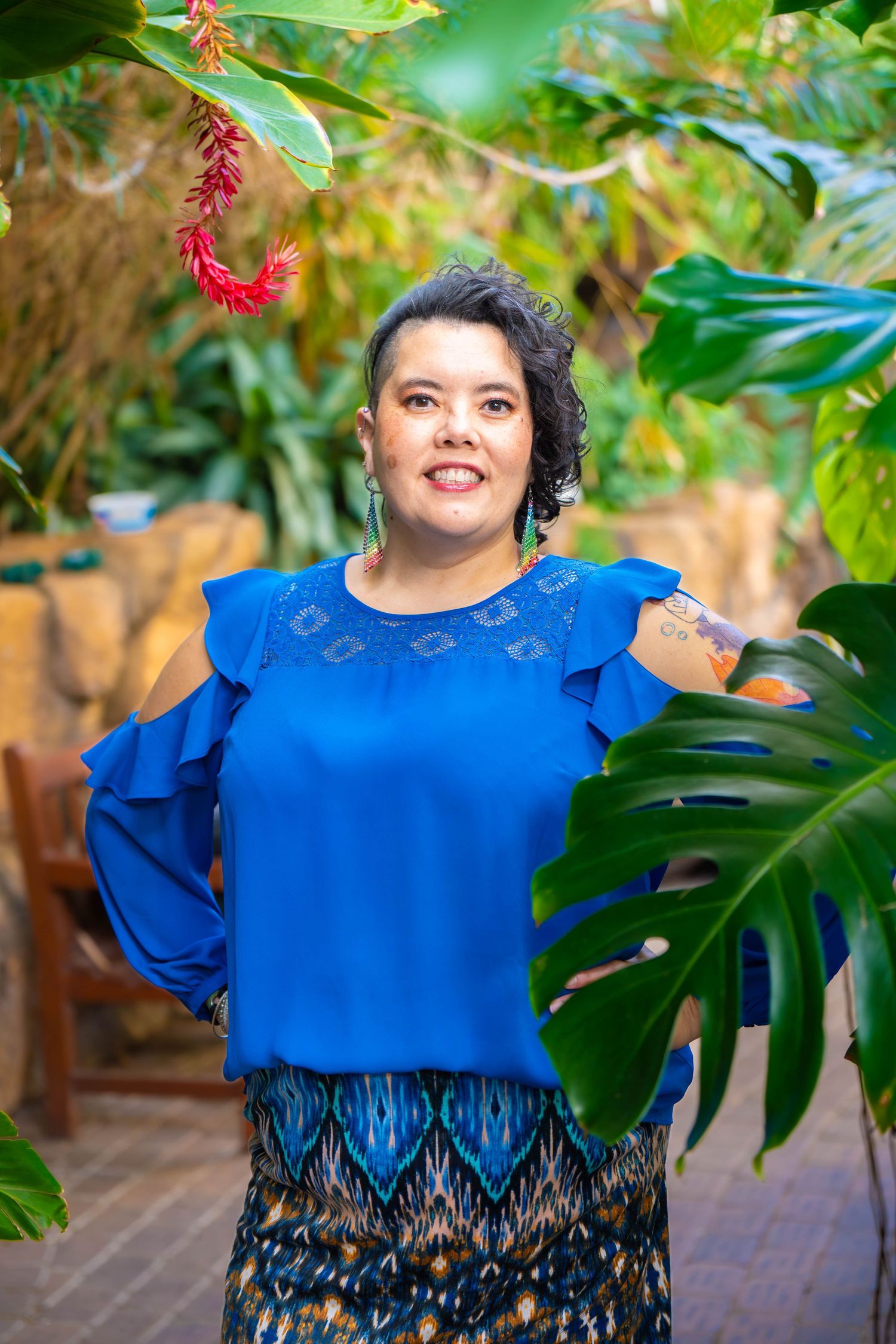 A woman in a vibrant royal blue top with ruffled sleeves poses outdoors against a lush green garden background.