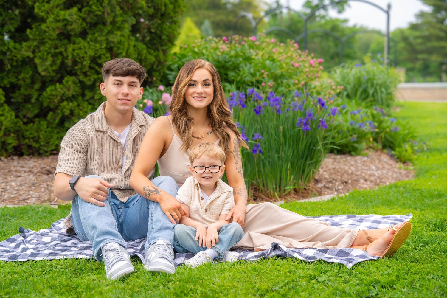 Young family sitting on blanket in garden surrounded by vibrant purple irises and lush greenery. Family Quad Cities Photography