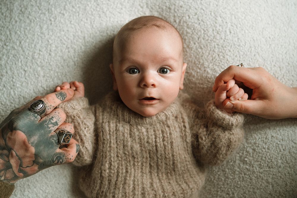Newborn photography in Manchester. Baby wearing a cream knit sweater lies on a textured grey surface while hands hold their tiny fingers.