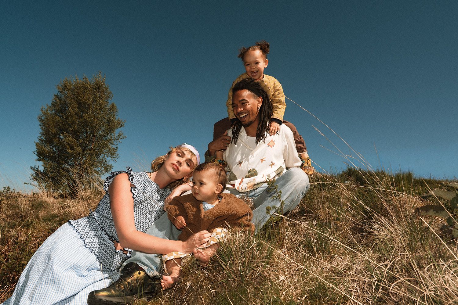 Two mothers and their children laugh and play together on a grassy hillside under a bright blue sky.
