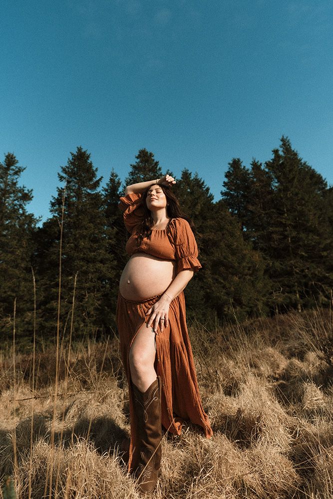 Pregnant woman in rust brown dress posing outdoors in field with pine trees and blue sky background.