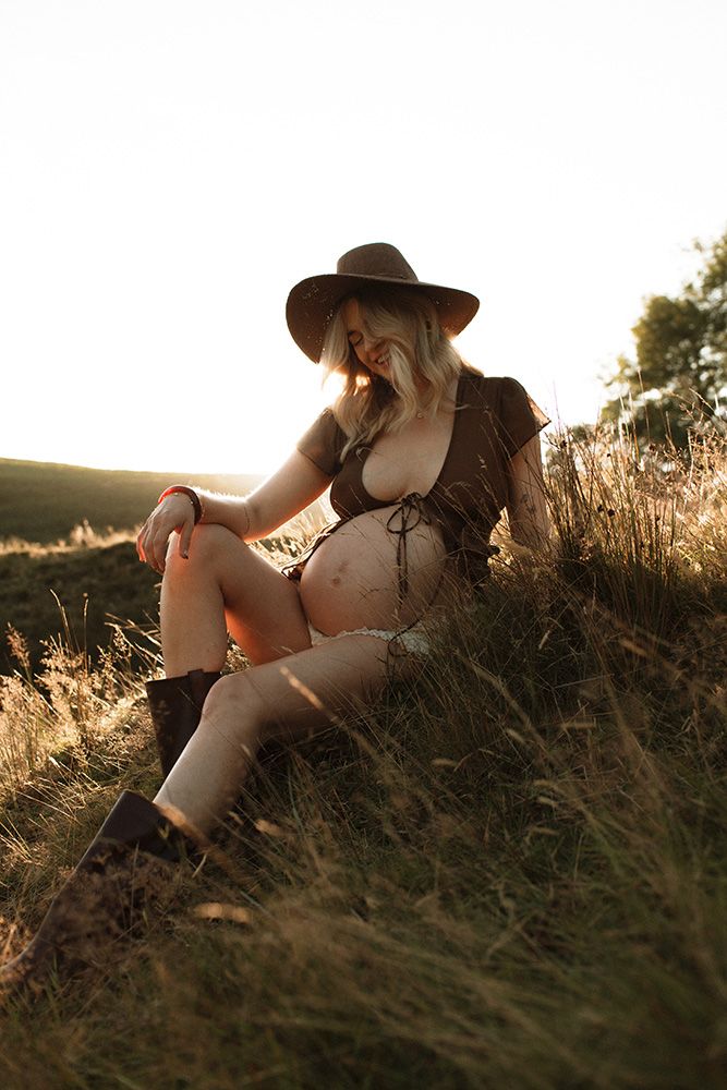 Lancashire maternity photographer. Silhouetted figure in a hat lying in golden backlit grass during sunset in a dreamy summer scene.