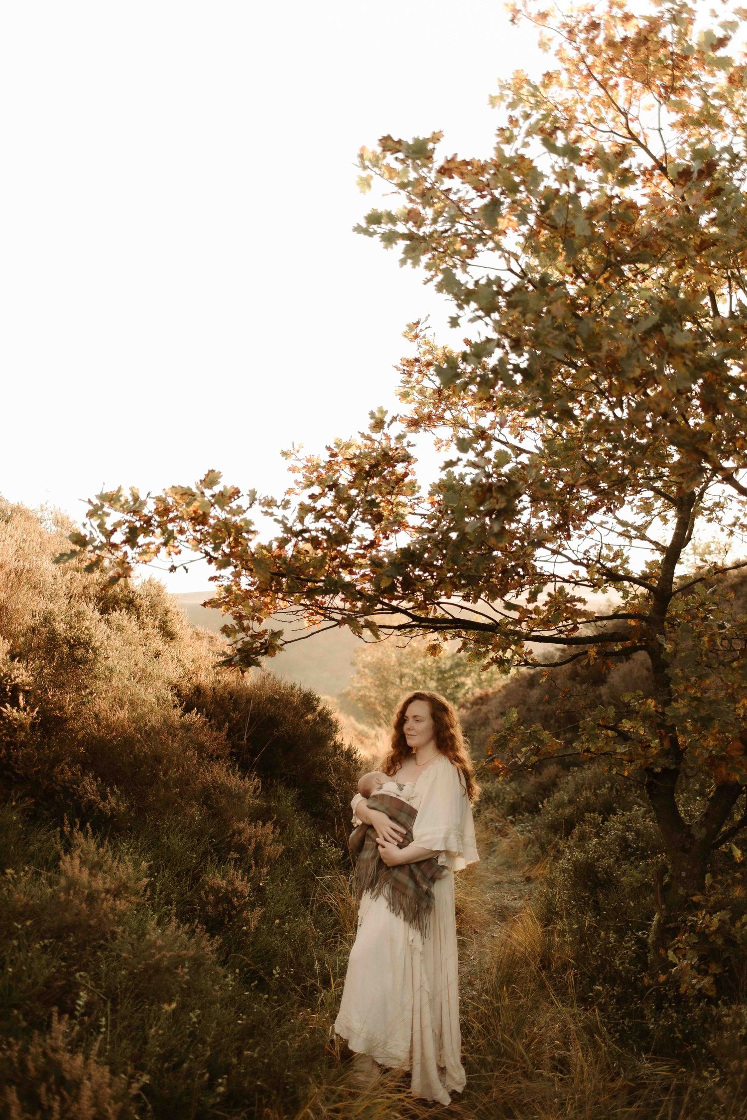 A person in a flowing white dress stands on a hillside path at golden hour beneath an autumn tree.