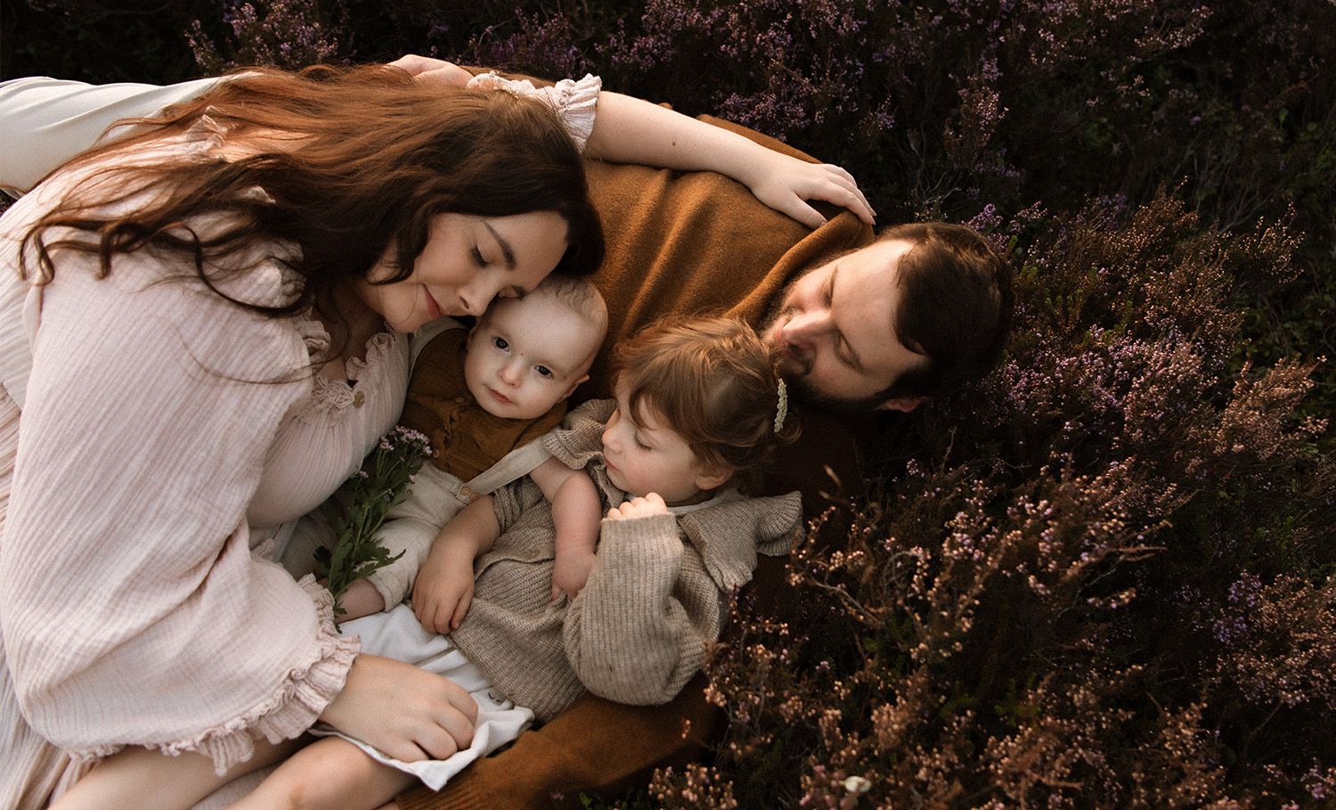 Family in light coloured clothing embraces on ground surrounded by autumn foliage in moody outdoor family photoshoot portrait, Chorley