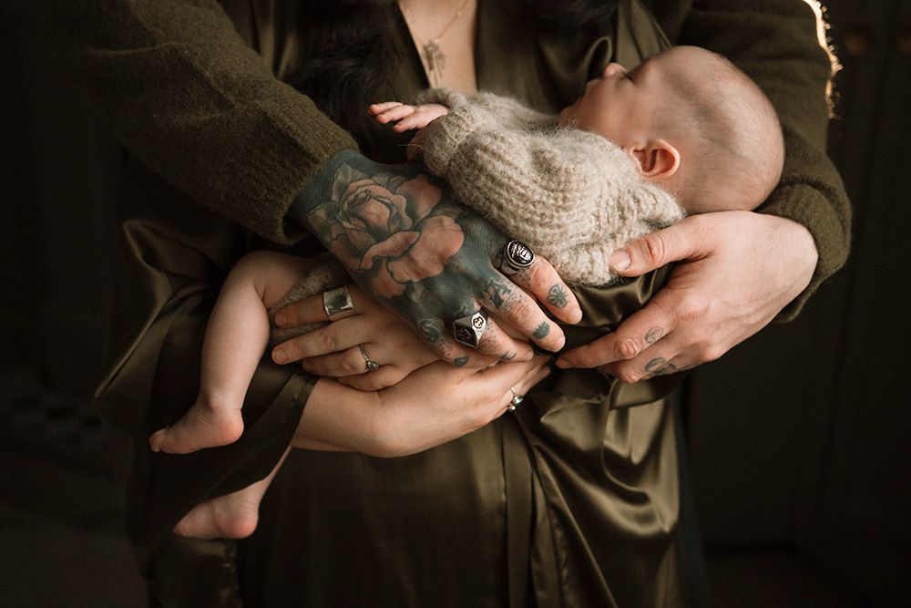 Parents cradling newborn baby in tattooed and ringed hands, wearing olive green clothing in soft light.