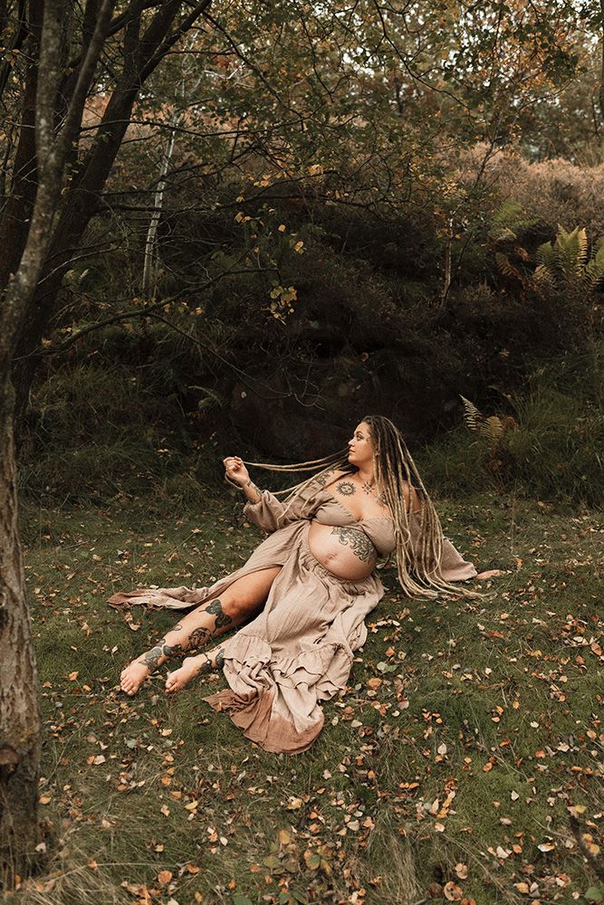 A pregnant woman with dreads in a boho dress sits among autumn leaves in a lush forest, during a natural maternity photoshoot in Manchester