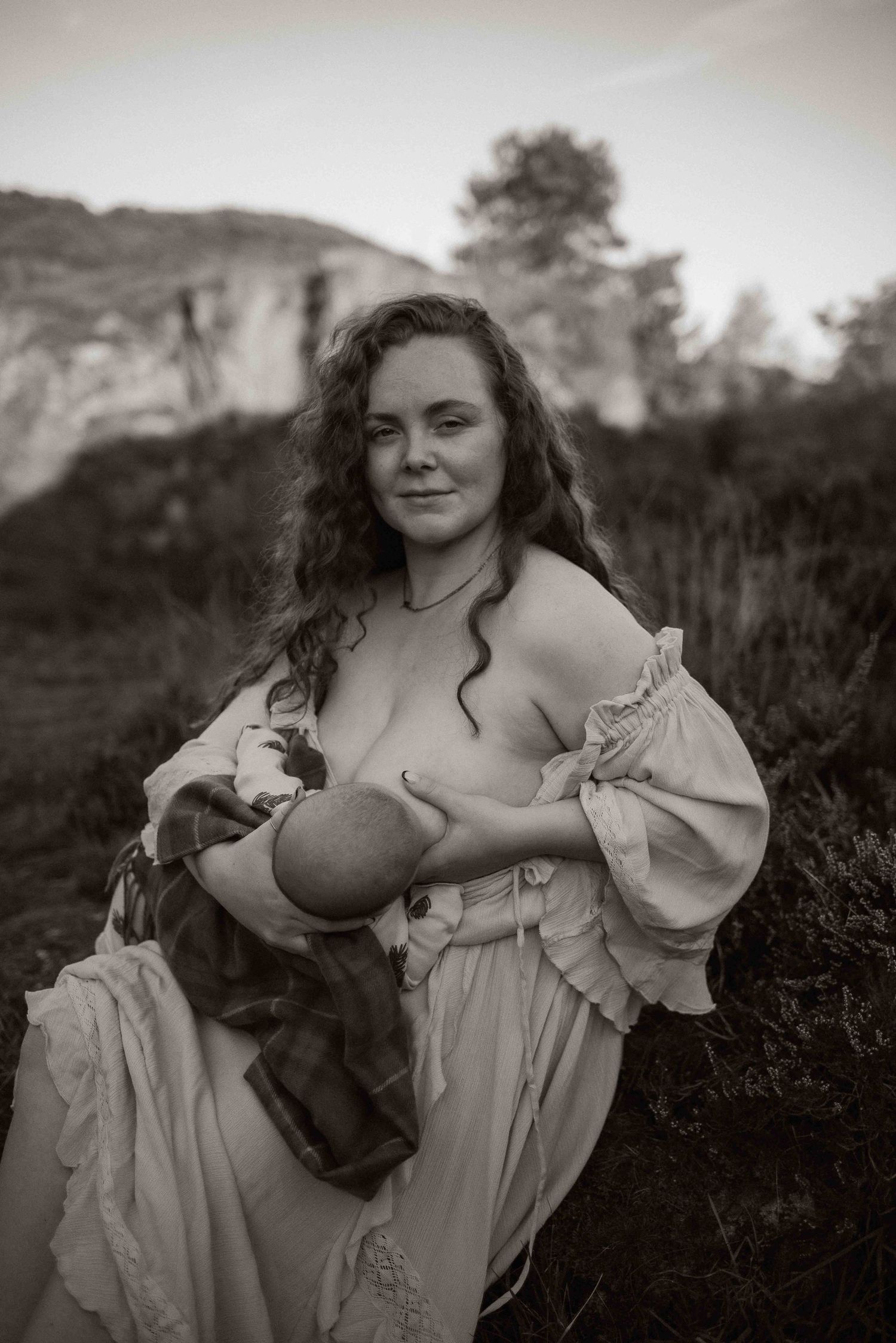 Black and white portrait of a nursing parent in a flowing dress outdoors with mountains in the background.