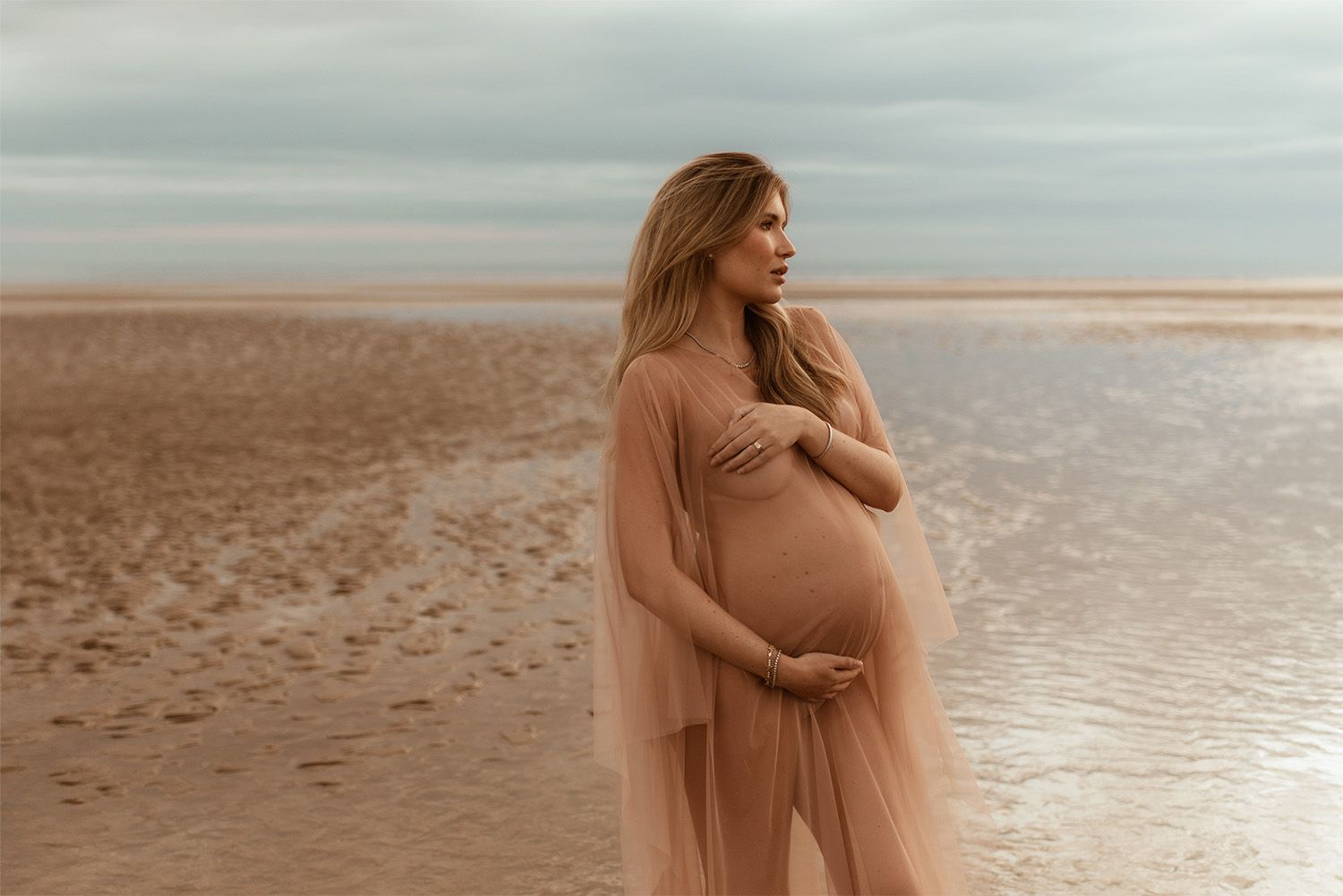 Pregnant woman in flowing peach dress stands on Formby beach at sunset holding her pregnant belly during maternity photoshoot