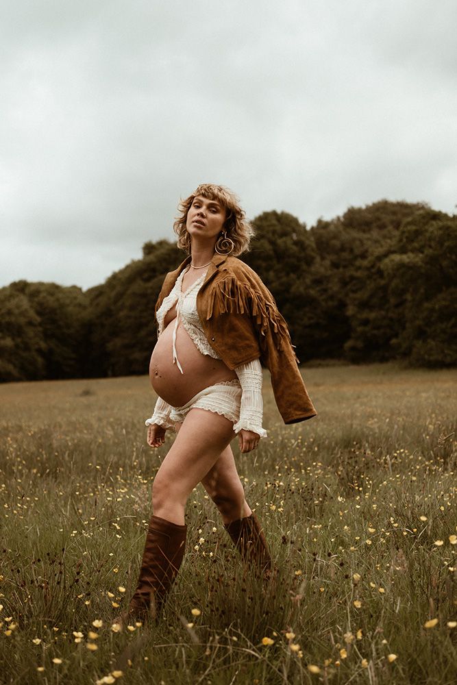 A pregnant woman wearing fringe leather jacket & boots stands in a wildflower meadow during golden hour, maternity photoshoot in Lancashire.