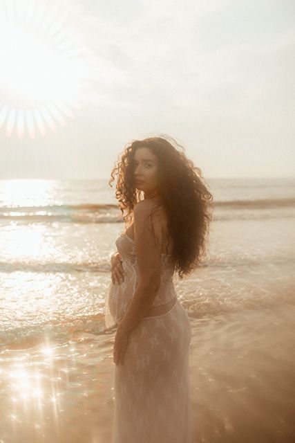 Lancashire maternity photographer. A pregnant Mum in a white lace dress stands on a beach with curly hair illuminated by golden light.