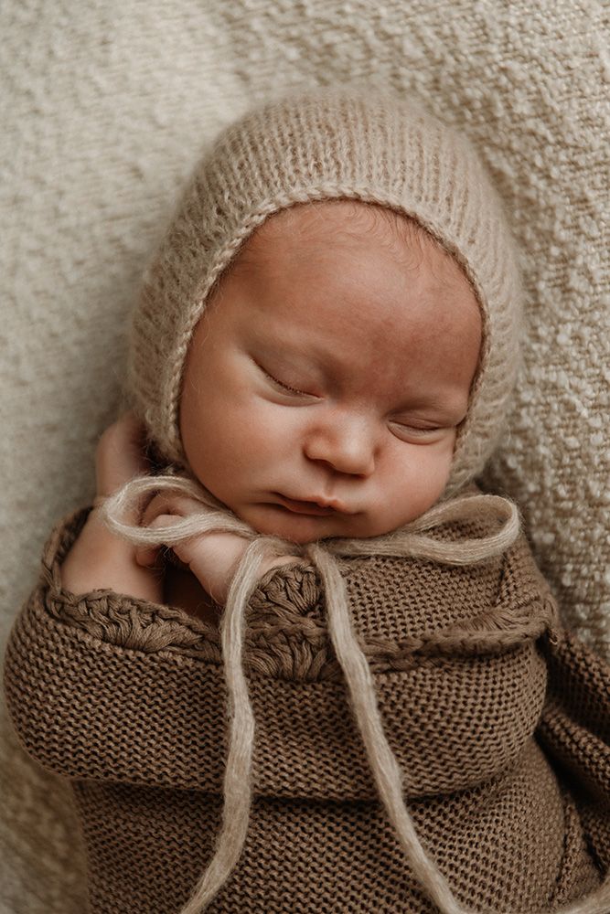 A sleeping newborn wrapped in a brown knit blanket with a cream colored bonnet rests peacefully for newborn photography in Lancashire.