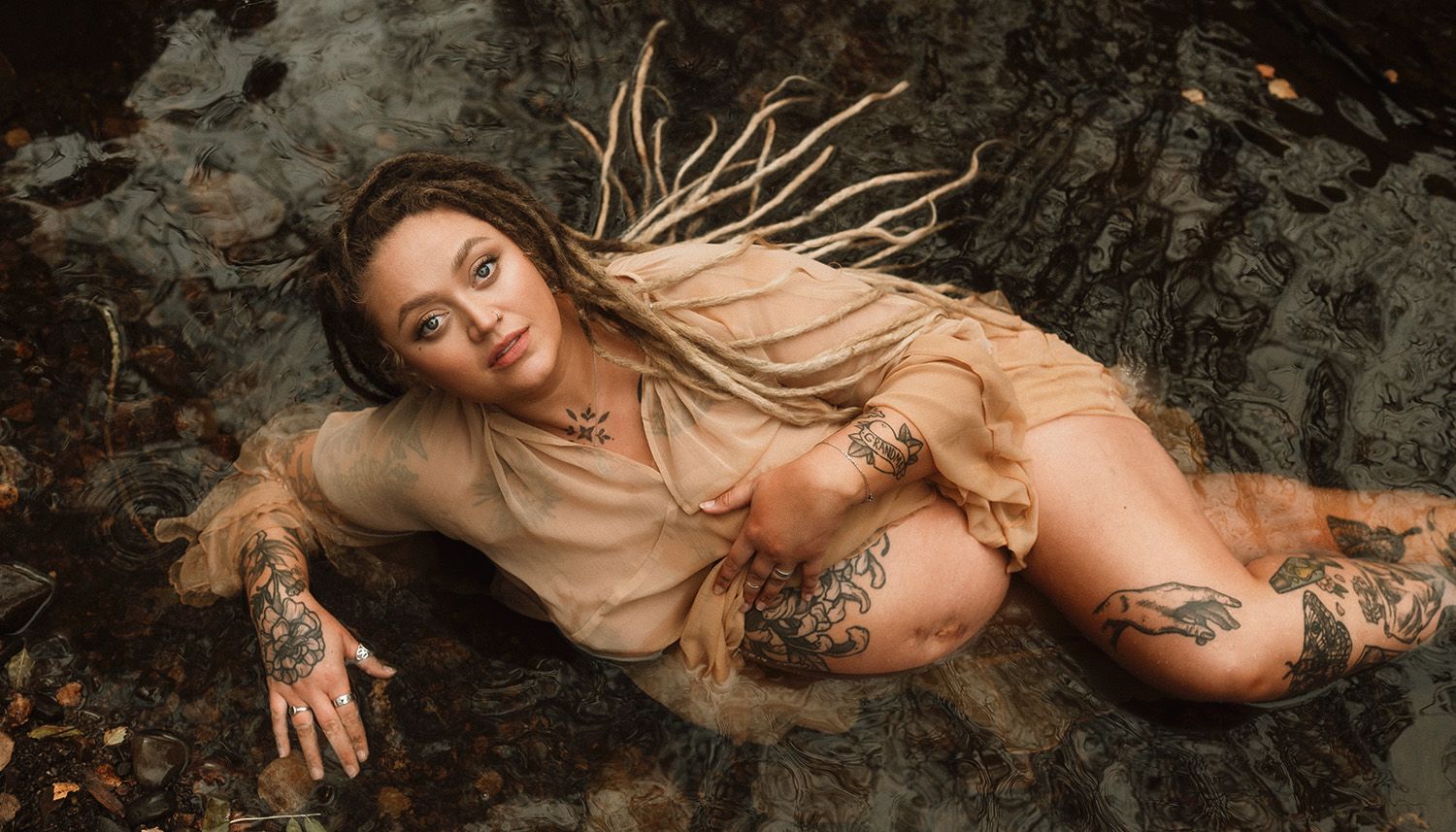 A moody maternity photoshoot in Lancashire featuring artistic poses of a tattooed mother in a rocky pool of water in natural lighting in.