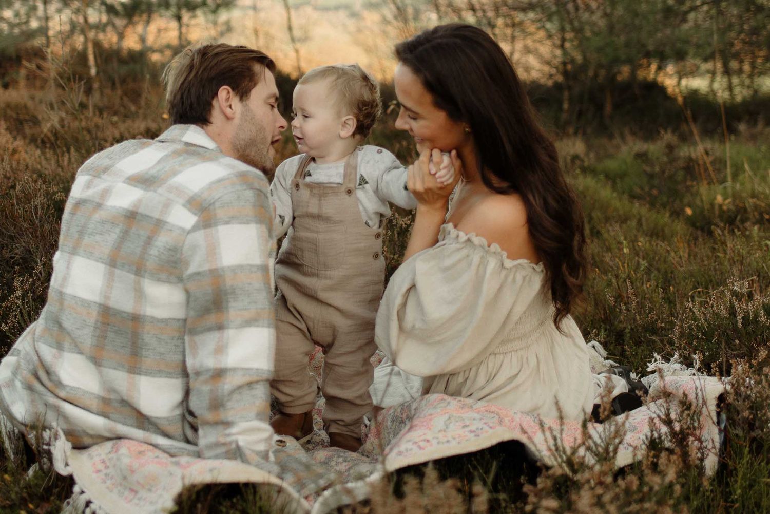 A family sits together on a blanket outdoors during a golden hour photoshoot in a rustic field setting.
