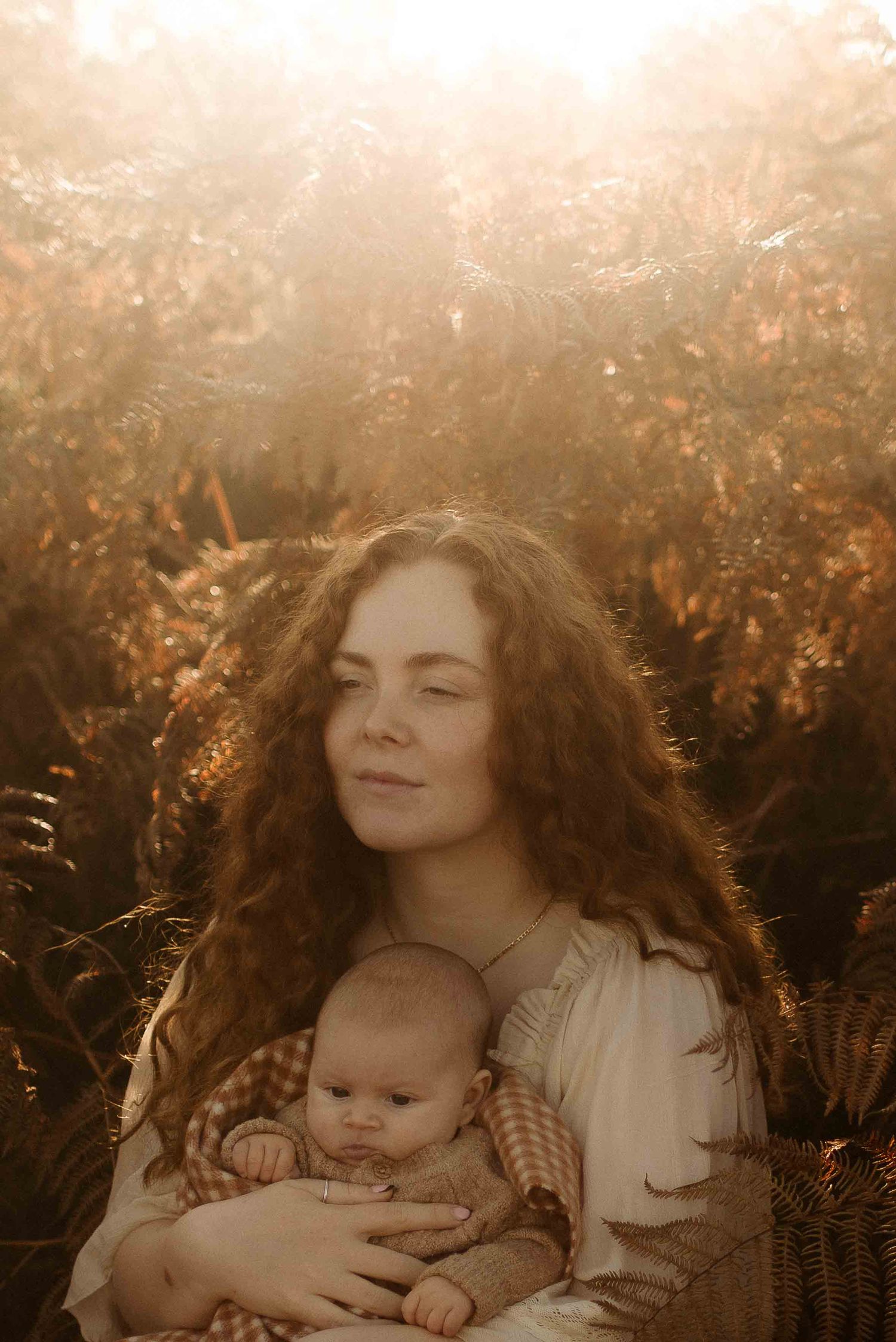 Ethereal nature photograph of mother and baby embracing in golden sunlit field with ferns during sunset.