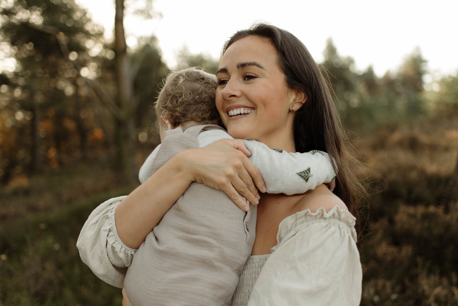 A person in a white blouse hugs a young child while smiling warmly in a sunlit outdoor setting during autumn.