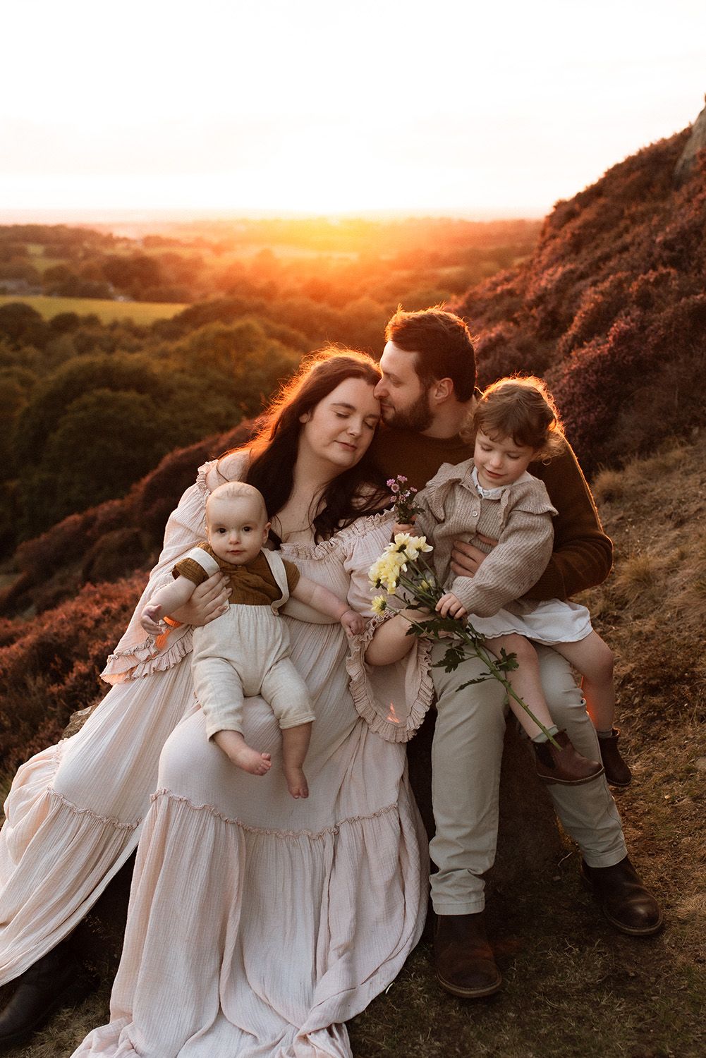 Children in light colored outfits play and embrace on a hilltop during golden hour sunset family photoshoot in Lancashire.