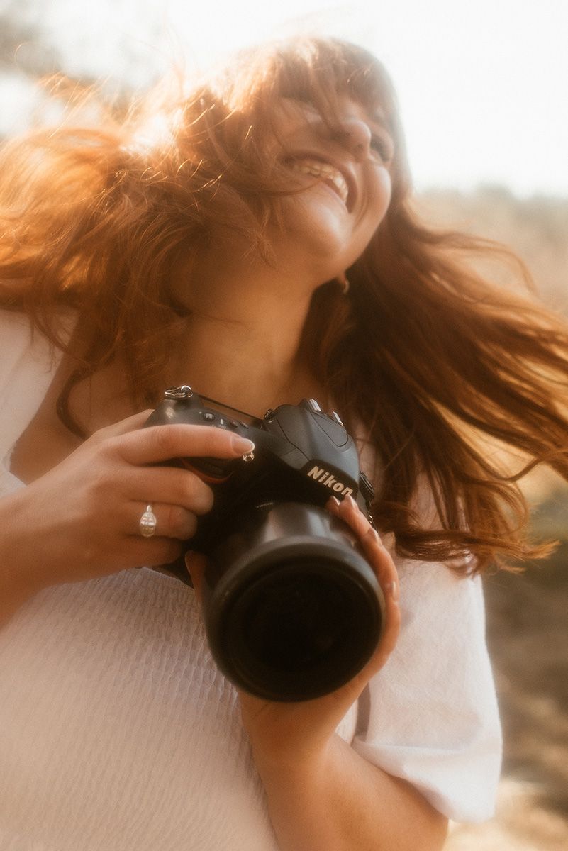 Photographer in white sweater holding camera with long lens while hair blows in golden sunlight.