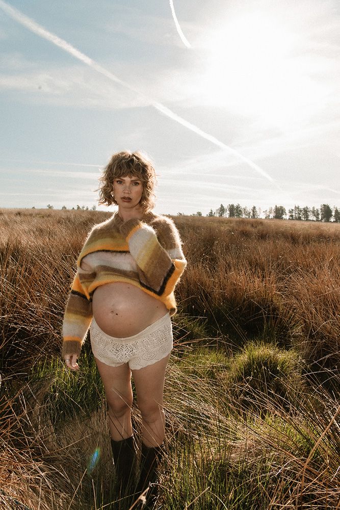 Pregnant woman in striped sweater and lace shorts stands in golden field under sunny sky with contrails. Pregnancy photoshoot in Manchester.
