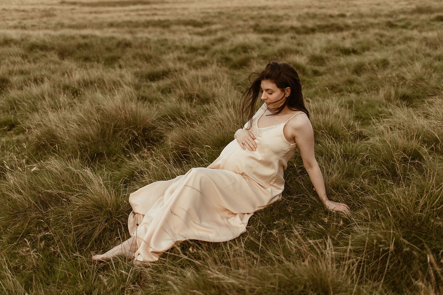 A pregnant mother in a flowing cream dress reclines in tall grass during a romantic outdoor maternity photoshoot.