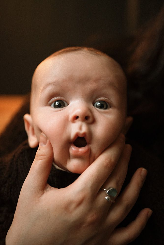 Newborn photography in Manchester. A surprised baby with wide eyes and open mouth being held by hands wearing a silver ring.