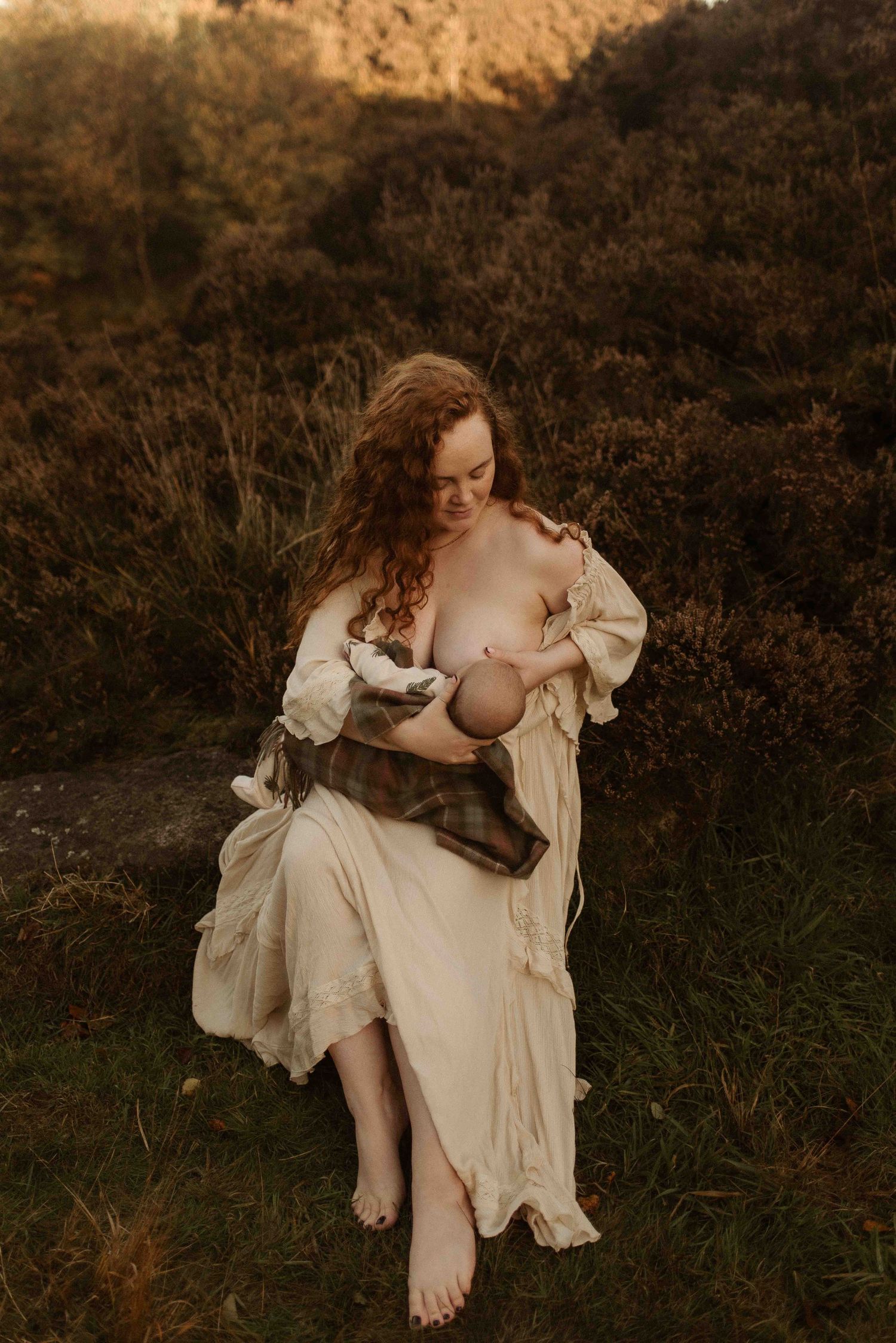 An ethereal figure in a white flowing dress sits barefoot on dark grassy ground at golden hour.