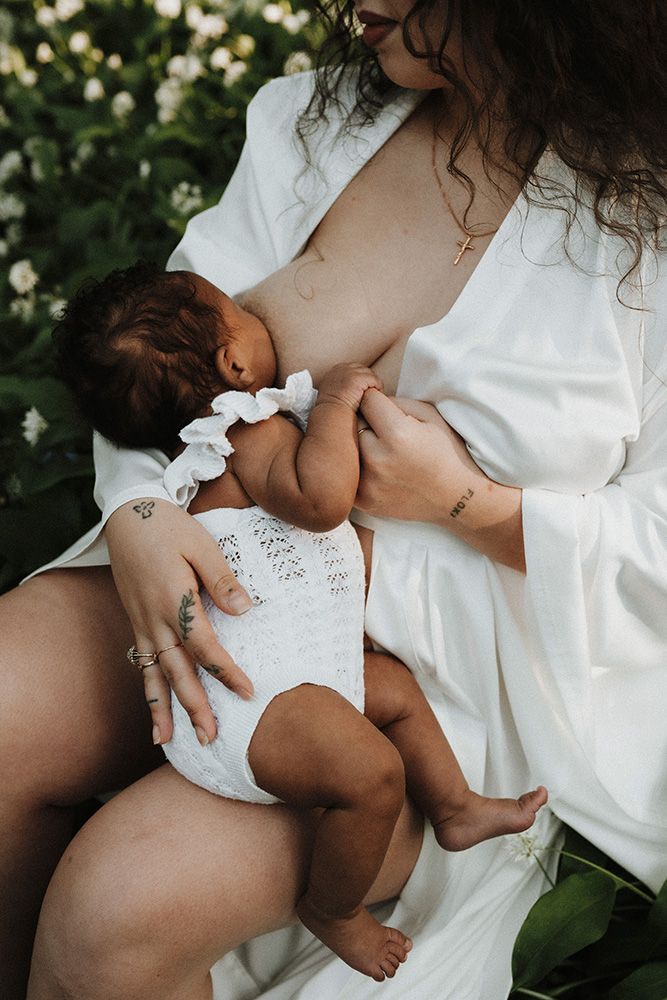 Mother in white robe breastfeeding newborn baby outdoors among green foliage and white flowers.