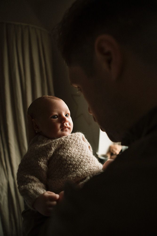 A dark, moody photograph showing a baby in white clothing against a shadowy background. Newborn photography at home in Manchester.