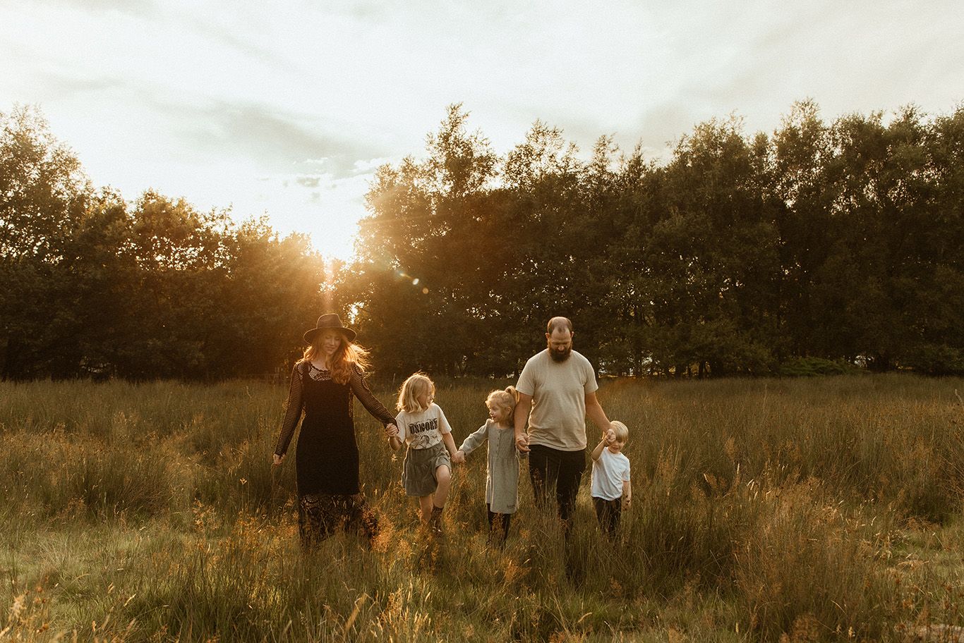 Family walks together through a golden field at sunset as sunlight streams through trees at White Coppice in Chorley, family photoshoot.