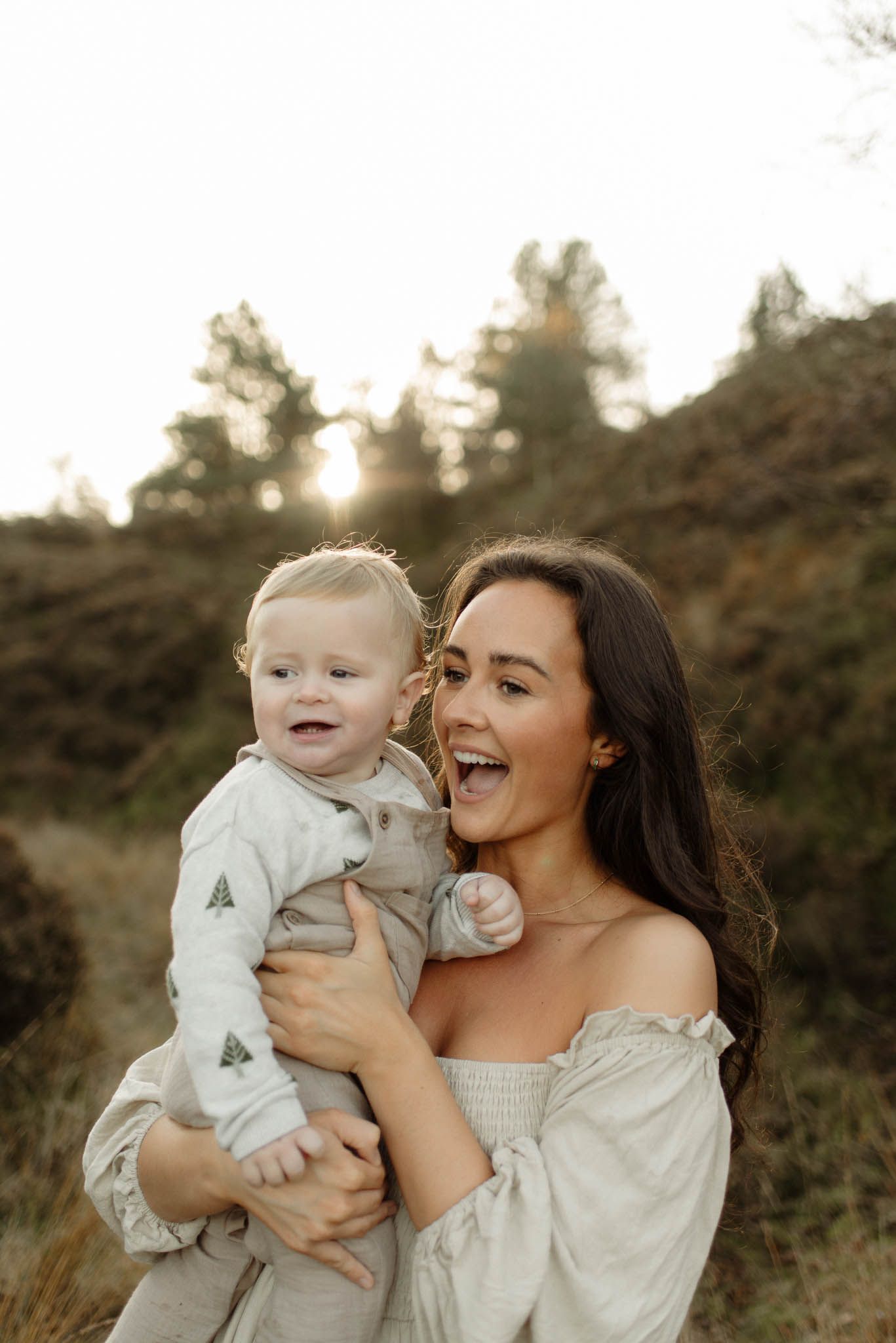 A candid portrait of a woman in a white off-shoulder dress holding a toddler in soft afternoon light.