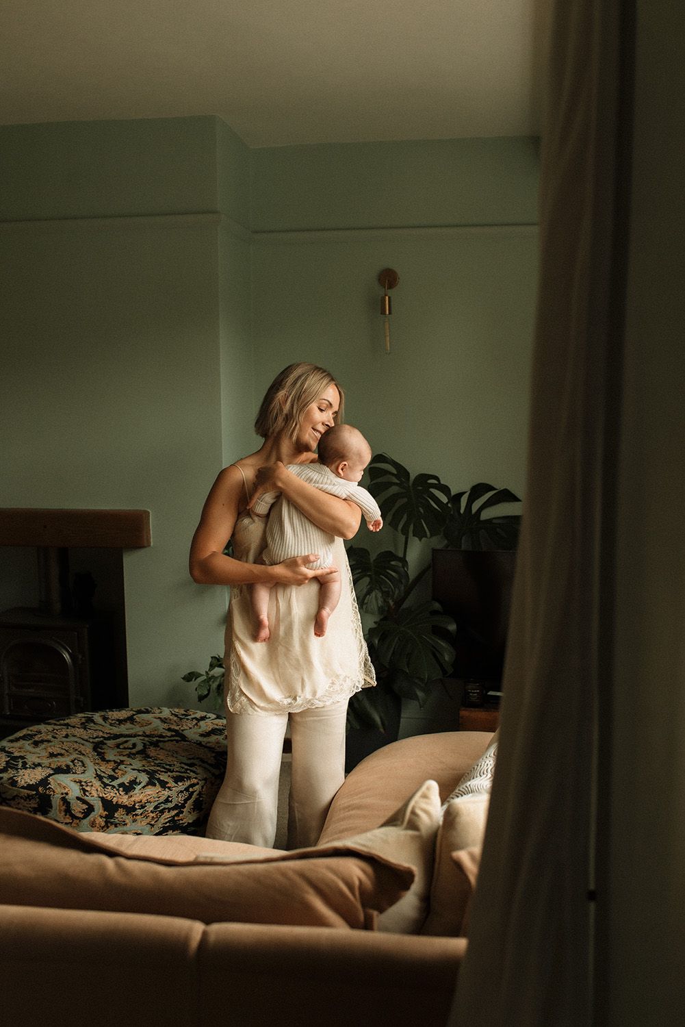 Two people in white clothing embrace in a dimly lit room with houseplants visible in the background, on a family photoshoot in Lancashire.