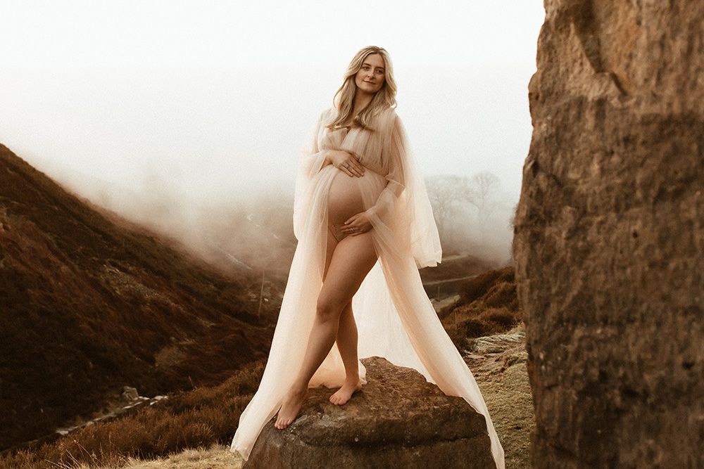 Pregnant woman in flowing sheer gown poses on rocky misty mountain landscape for maternity photography in Manchester session.